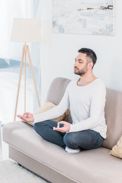 przystojny mężczyzna z oczami zamknięte siedzi na kanapie w Lotus Pose i medytowanie w domu - Zdjęcie, obraz