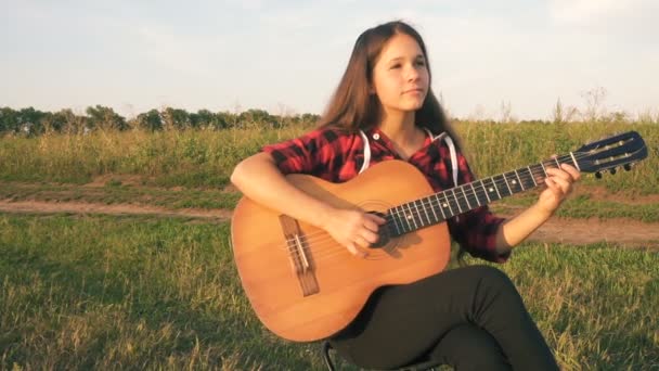 Young girl playing guitar on meadow at the sunset - Кадры, видео