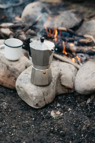 Closeup photo of white cup and coffee maker near to bonfire. Con - Фото, изображение