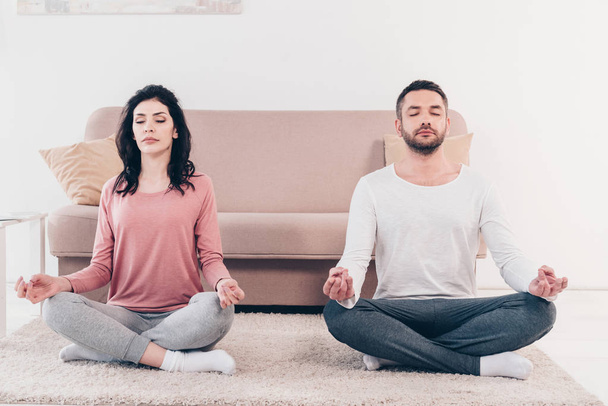 Frau und Mann mit geschlossenen Augen sitzen in Lotuspose und meditieren zu Hause - Foto, Bild