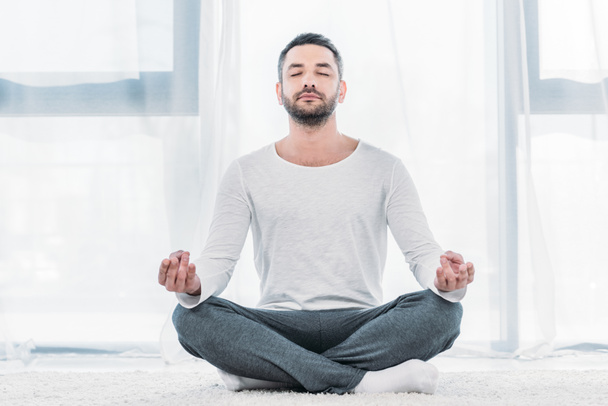 gutaussehender Mann mit geschlossenen Augen sitzt auf Teppich in Lotus-Pose und meditiert zu Hause - Foto, Bild