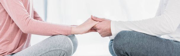 panoramic shot of man and woman sitting in lotus pose, meditating and holding hands at home - Photo, image