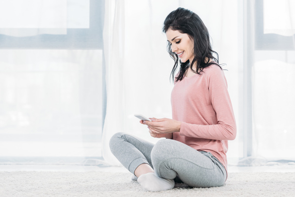 beautiful smiling woman sitting on carpet and using smartphone at home with copy space - Foto, afbeelding