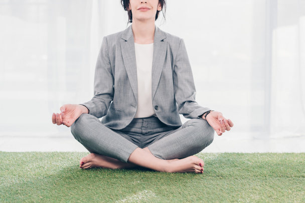 Vista recortada de la mujer de negocios en traje meditando mientras está sentado en la estera de hierba en Lotus Pose
  - Foto, imagen