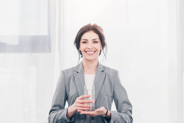 beautiful smiling businesswoman in suit holding glass of water and looking at camera - Zdjęcie, obraz