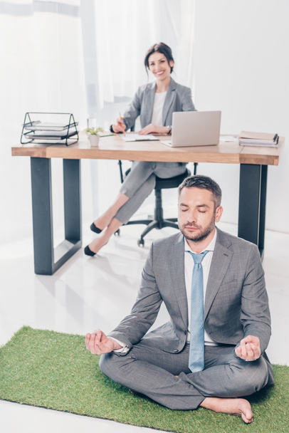 businessman meditating in Lotus Pose on grass mat while businesswoman sitting at table in office - Фото, изображение