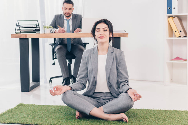 empresária meditando em Lotus Pose no tapete de grama, enquanto empresário sentado à mesa e escrevendo na mesa no escritório
 - Foto, Imagem
