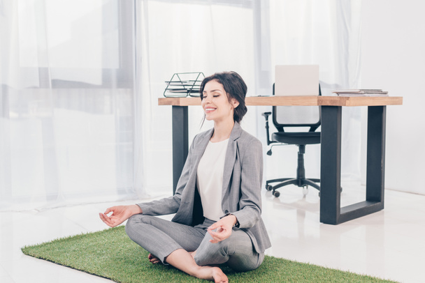belle femme d'affaires heureuse en costume assis sur tapis d'herbe dans Lotus Pose et méditer dans le bureau
 - Photo, image