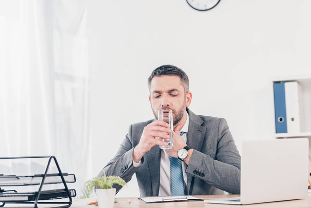 schöner Geschäftsmann im Anzug, der am Schreibtisch sitzt, die Kehle berührt und im Büro Wasser trinkt - Foto, Bild