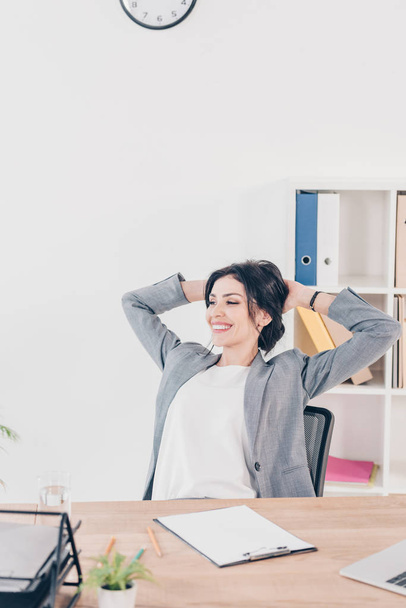 beautiful smiling businesswoman in suit with hands on head sitting at table in office  - 写真・画像