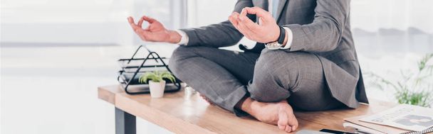 Panoramaaufnahme eines Geschäftsmannes, der in Lotus-Pose auf seinem Schreibtisch meditiert - Foto, Bild