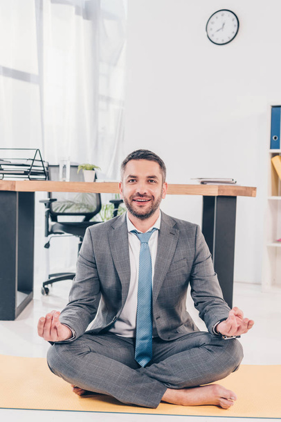 przystojny uśmiechnięty biznesmen medytacja w Lotus Pose na mat fitness w biurze - Zdjęcie, obraz