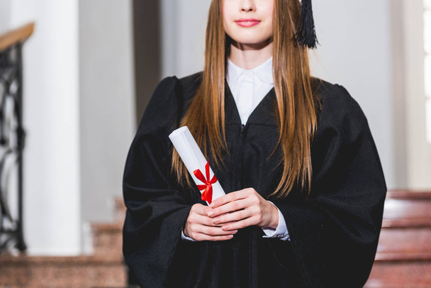 oříznutý pohled na veselou mladou ženu, která drží diplom a usmívá se na univerzitě  - Fotografie, Obrázek