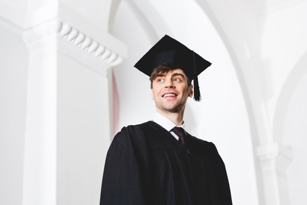 vue à angle bas du jeune homme heureux dans le chapeau de graduation souriant à l'université
  - Photo, image