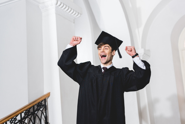 happy young man in graduation cap smiling and gesturing in university  - 写真・画像