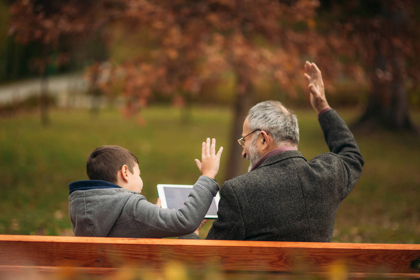 El abuelo y el nieto se sientan en el banco y usan una tableta. Levantaron la mano.
 - Foto, Imagen