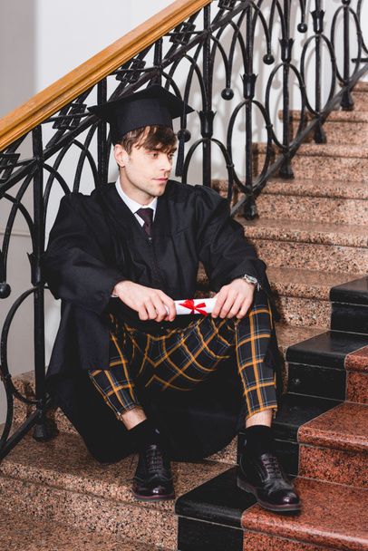 mezuniyet elbisesi içinde yakışıklı öğrenci merdivenlerde oturan ve diploma tutan  - Fotoğraf, Görsel