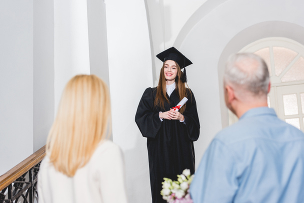 selective focus of cheerful girl in graduation cap looking at parents and holding diploma  - Foto, Imagem