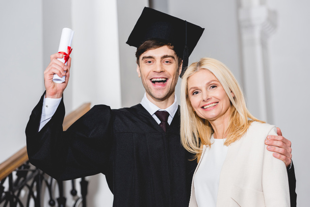 cheerful son in graduation cap holding diploma while standing with mother  - Фото, зображення