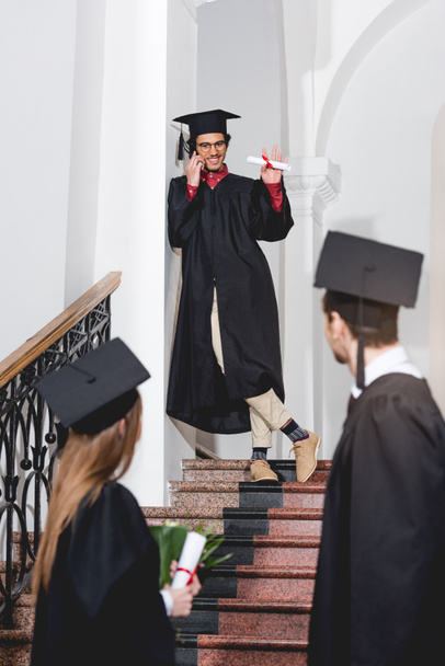 cheerful man talking on smartphone and waving hand to students in graduation caps  - Photo, Image