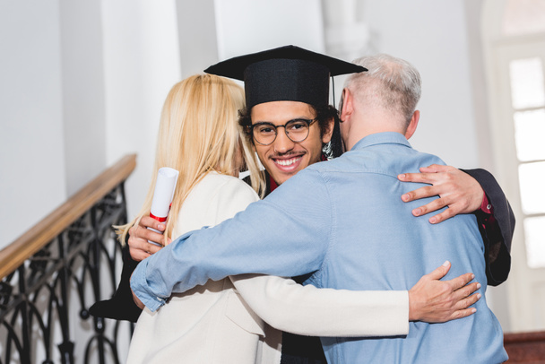 Вид сзади зрелых родителей, обнимающих счастливого сына в очках и выпускной шапке
 - Фото, изображение