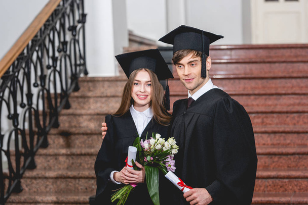 przystojny mężczyzna i atrakcyjna dziewczyna z kwiatami uśmiechniętymi podczas posiadania dyplomów  - Zdjęcie, obraz