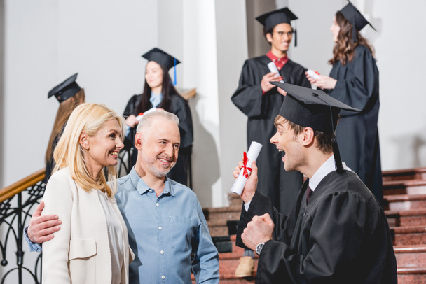 selective focus of cheerful parents looking at happy son in graduation cap gesturing while holding diploma near students  - Photo, Image