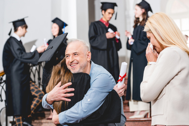 back view of girl in graduation cap hugging with father near crying mother - Photo, Image