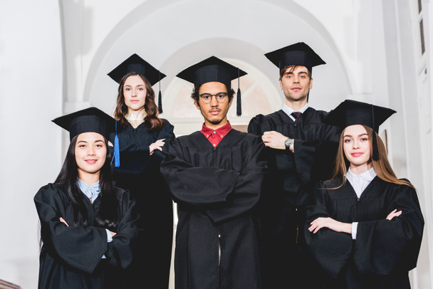 estudantes alegres em vestidos de graduação de pé com braços cruzados na universidade
  - Foto, Imagem