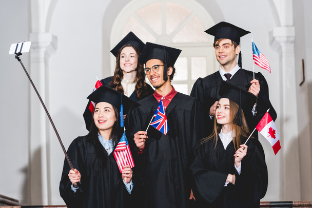 vrolijke studenten in afstuderen jurken houden vlaggen van verschillende landen en het nemen van selfie op smartphone  - Foto, afbeelding
