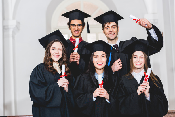 glückliche Gruppe von Studenten in Abschlusskleidern, die lächelnd Diplome in der Hand halten  - Foto, Bild