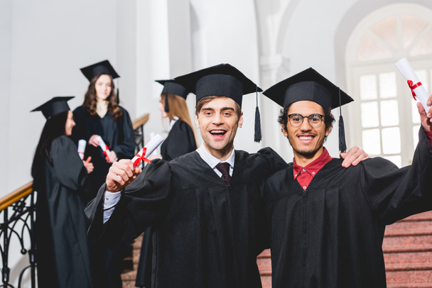 Selektiver Fokus fröhlicher Männer in Abschlussmützen, die Diplome in der Nähe von Studenten halten  - Foto, Bild
