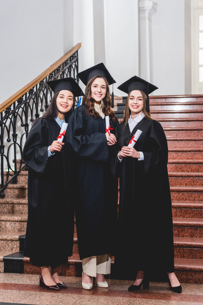 estudiantes alegres y bonitos en gorras de graduación sonriendo mientras sostiene diplomas en la universidad
  - Foto, imagen