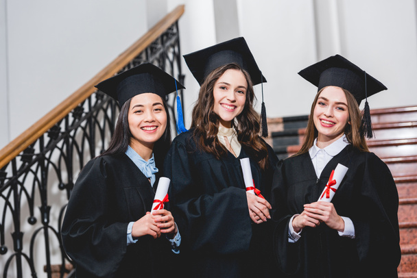 cheerful and beautiful students in graduation caps smiling while holding diplomas in university  - Photo, Image