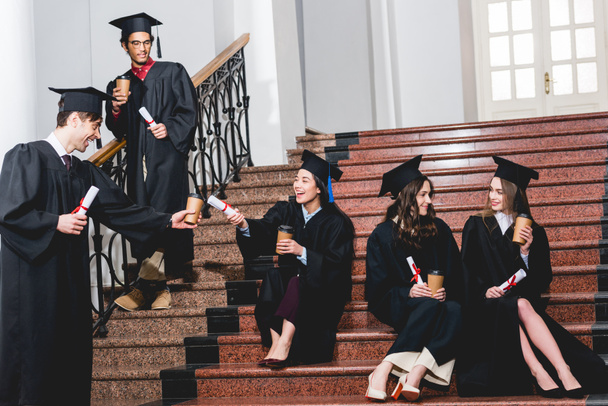 vidám csoport hallgatók érettségi ruhák lépcsőn ülve, papírpoharak és diplomák  - Fotó, kép