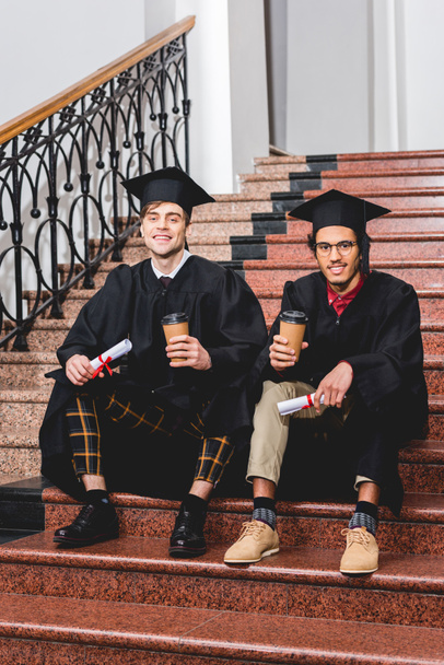 estudiantes alegres en vestidos de graduación con diplomas y vasos de papel mientras están sentados en las escaleras
  - Foto, Imagen