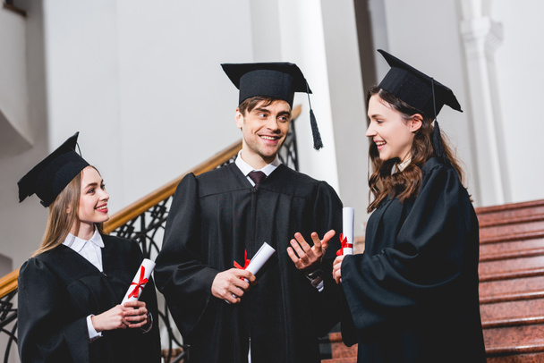 cheerful man in graduation cap gesturing near attractive girls while holding diploma  - Fotoğraf, Görsel