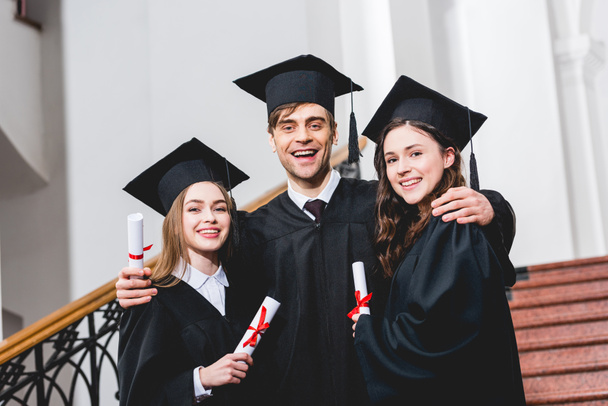 cheerful man in graduation cap hugging attractive girls while holding diploma  - Photo, Image