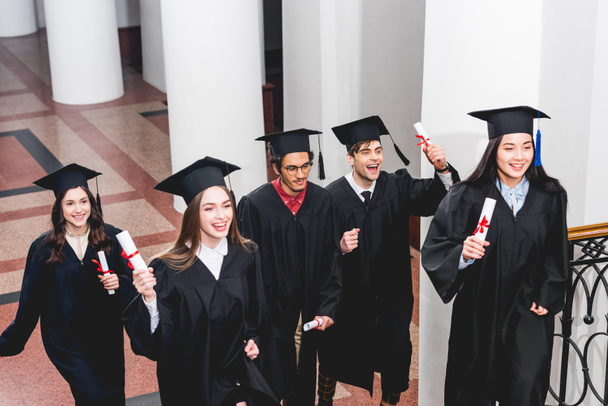 estudantes sorridentes em vestidos de graduação titulares de diplomas na universidade
  - Foto, Imagem