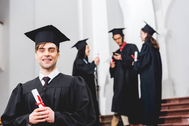foco seletivo do homem feliz em vestido de graduação olhando para câmera e segurando diploma perto de estudantes
  - Foto, Imagem