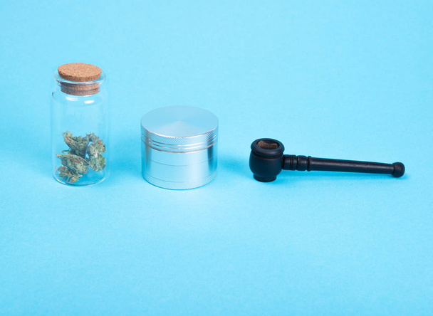 Dried medical cannabis buds in a glass bottle, silver grinder and wooden pipe on blue background.  - Фото, зображення