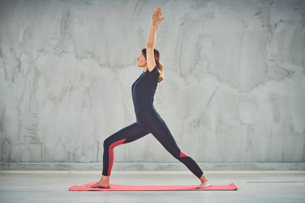 Vista laterale della brunnette Caucasain in piedi sul tappeto in posizione Crescent Lunge yoga
. - Foto, immagini
