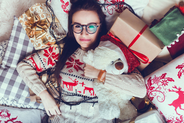 Una hermosa chica en un suéter de punto con ciervos y un cuadros cumple con la Navidad y un nuevo año, examina y da regalos, sueños, un ambiente festivo y ambiente, utilizando teléfono inteligente, cara feliz, sonrisa, hogar
 - Foto, imagen