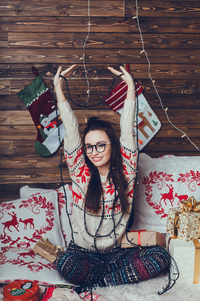 Una hermosa chica en un suéter de punto con ciervos y un cuadros cumple con la Navidad y un nuevo año, examina y da regalos, sueños, un ambiente festivo y ambiente, utilizando teléfono inteligente, cara feliz, sonrisa, hogar
 - Foto, imagen