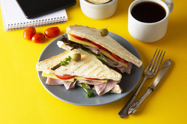 Snacks, fast food concept. Eatting at work place. Fresh club sandwich, vegetables, coffee, potato chips, sweet cookies. Tablet screen. Yellow background. - Φωτογραφία, εικόνα