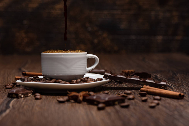 Pouring hot black coffee from cezve into the cup. Coffee beans, chocolate, star anise on a wooden background. - 写真・画像