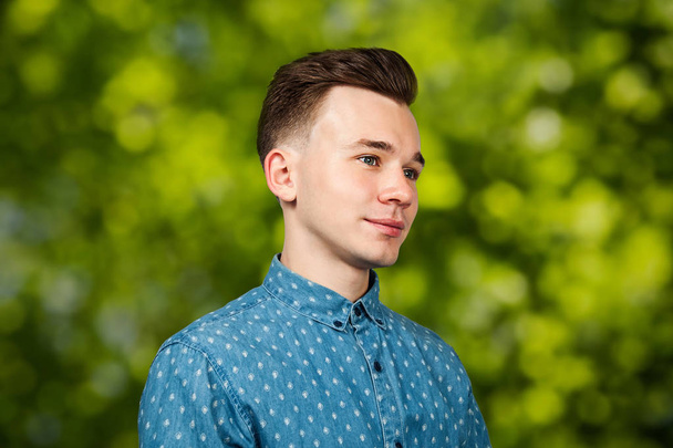 Profil lächelnd Porträt weiß junger Kerl Modell gekleidet in blauem Hemd auf grünem Bokeh Hintergrund - Foto, Bild