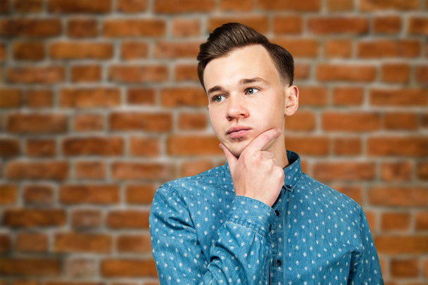 White young guy dressed in blue shirt holds hand behind head and looking aside on brick wall background - Фото, зображення