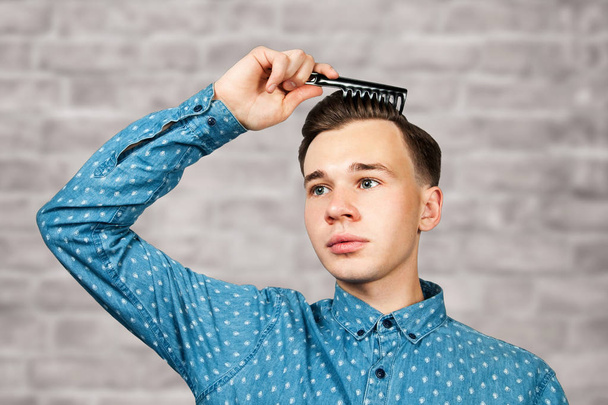 portrait white young man dressed in blue shirt. Man hold comb in his hair on brick wall background - Photo, Image