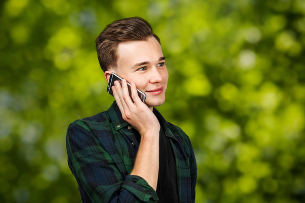 Valkoinen nuori mies kirjoittaa viestin matkapuhelimeen ja hymyilee. Mies vihreällä bokeh taustalla
 - Valokuva, kuva